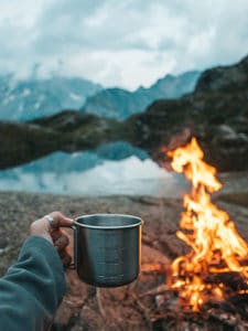 camper holding a mug by a fire and lake